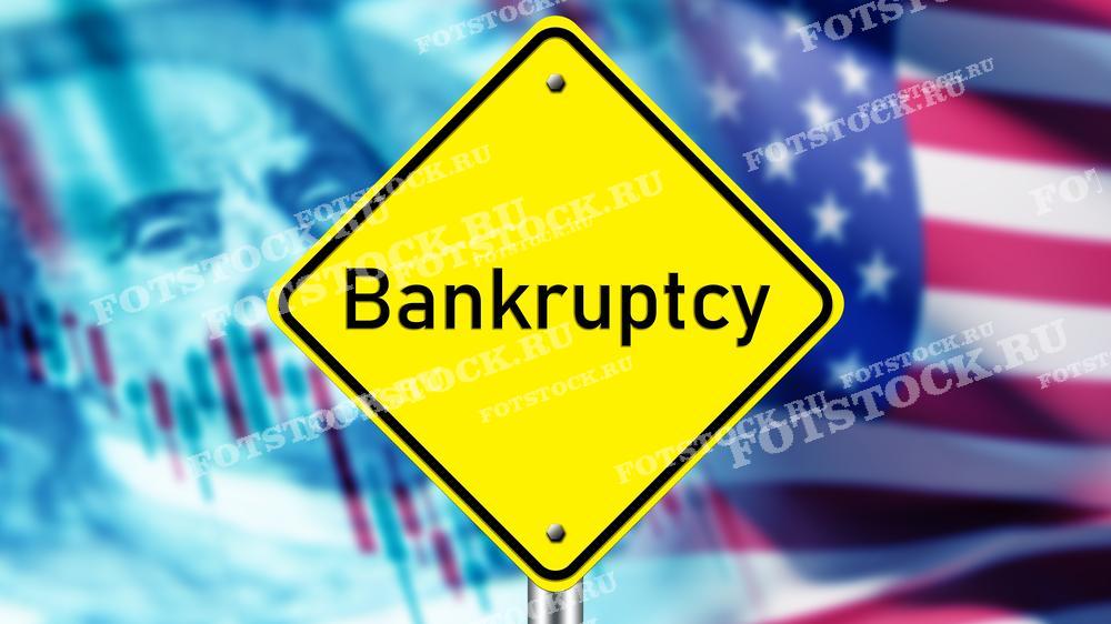 На фотографии изображен предупреждающий знак с надписью "Bankruptcy" на фоне американского флага и финансовых графиков. Снимок символизирует экономический кризис и банкротства в США, акцентируя внимание на финансовой нестабильности и экономических проблемах.