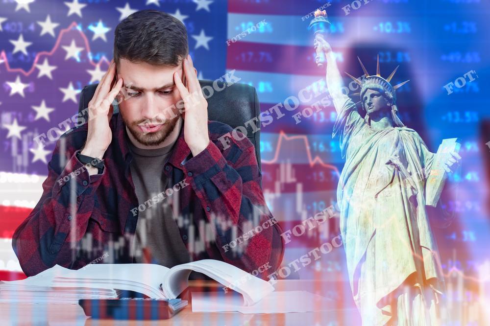 На изображении — обеспокоенный молодой человек с книгами, сидящий за столом, на фоне флага США, биржевых графиков и статуи Свободы. Фото символизирует финансовое давление, социальную нестабильность и вызовы, с которыми сталкиваются граждане в условиях экономических трудностей в США. Подходит для тем, связанных с образованием, стрессом, социальными проблемами и экономикой США.