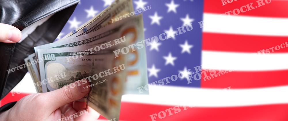Рука человека держит несколько стодолларовых банкнот на фоне флага США. Символизирует финансовую независимость, благосостояние и экономику Америки.