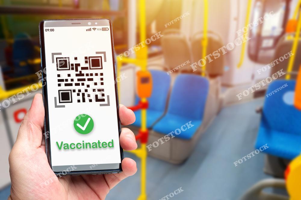 На фото изображена рука, держащая смартфон с QR-кодом и отметкой о вакцинации на экране. Фон составляют сиденья общественного транспорта, что символизирует требования предъявления доказательства вакцинации для поездок.