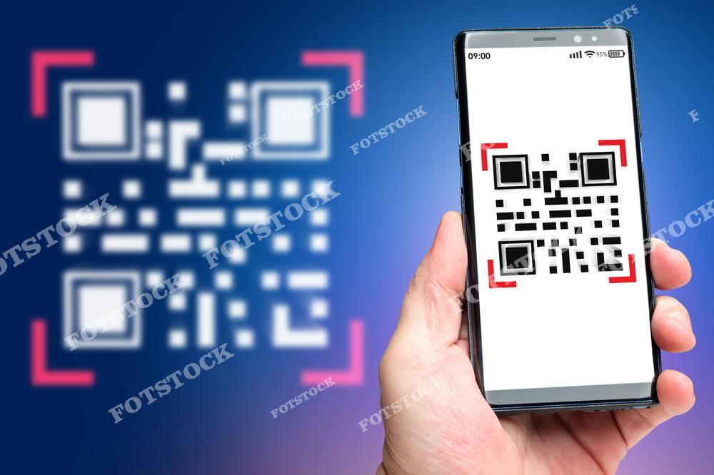 На фотографии изображена рука, держащая смартфон с отображением QR-кода на экране. На заднем фоне виден размытый крупный QR-код, что акцентирует внимание на технологии сканирования. Фото иллюстрирует удобство и актуальность цифровых технологий в повседневной жизни.