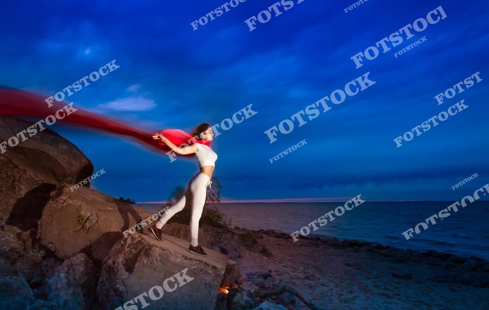 На фотографии изображена женщина в белом спортивном костюме, стоящая на скале у берега моря. Яркий красный шарф развивается за её спиной на фоне драматического синего неба. Образ передаёт динамику, внутреннюю силу и стремление вперёд.