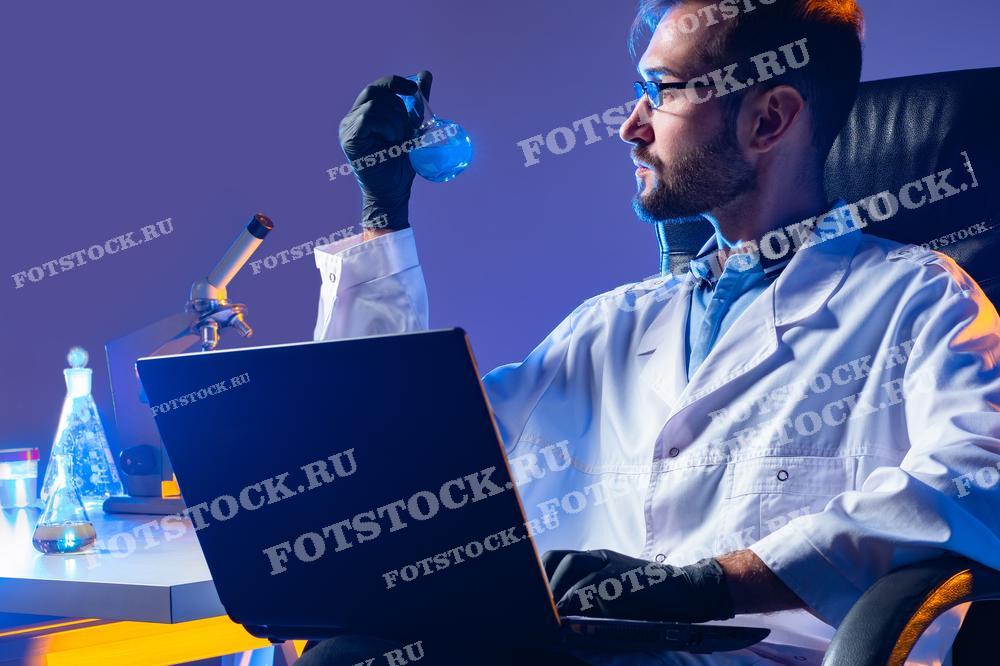 На фотографии изображен мужчина-ученый в лабораторном халате, работающий за столом с ноутбуком и микроскопом. Он держит колбу с синим раствором, внимательно рассматривая её содержимое. Фотография может быть использована для иллюстрации научных исследований, образовательных материалов, рекламных кампаний фармацевтических компаний или для изображения работы в лаборатории.