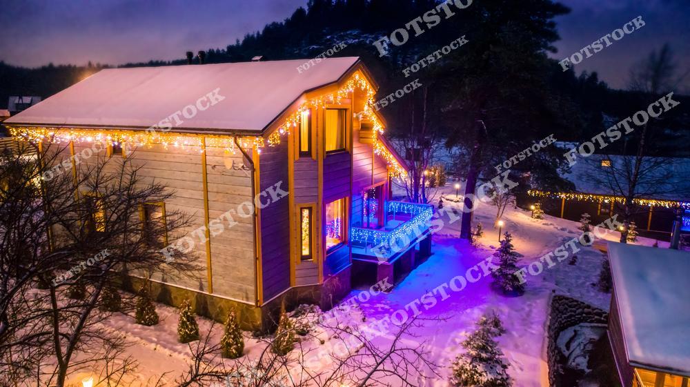 На фотографии изображен загородный дом в зимнем поселке, украшенный гирляндами, которые создают теплое освещение в вечернее время. Снег и праздничное освещение на террасе и вокруг дома подчеркивают атмосферу уюта и спокойствия. Изображение прекрасно передает зимнее настроение и дух новогодних праздников.