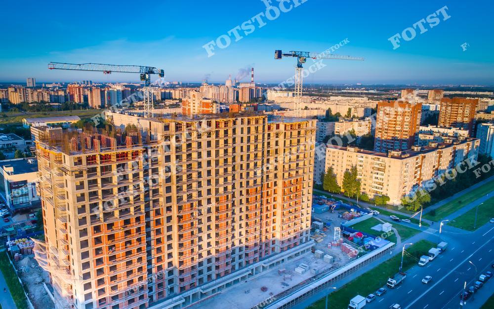 На фотографии изображен процесс строительства многоэтажного жилого комплекса с башенными кранами на фоне городской застройки. Здание находится на финальных этапах строительства, что символизирует развитие и рост городской инфраструктуры. Этот вид подчеркивает современный подход к строительству и архитектуре.