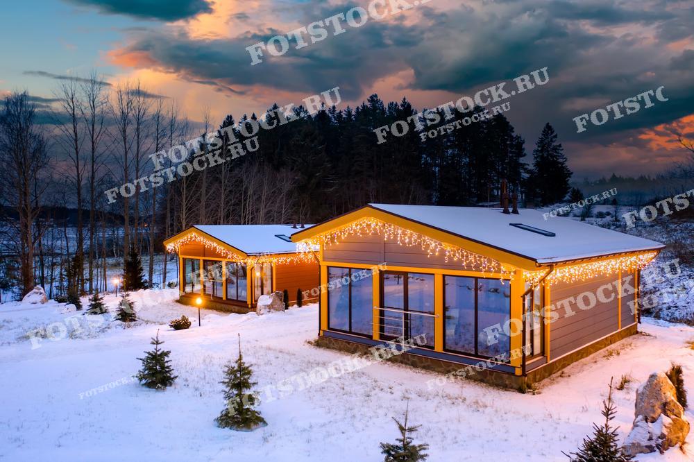 На фотографии изображены два загородных дома, украшенные светящимися новогодними гирляндами. Снег покрывает землю, создавая зимнюю атмосферу, а закатные краски неба добавляют тепла и уюта. Дома расположены среди деревьев, что подчеркивает уединенность и спокойствие этого места, создавая идеальные условия для зимнего отдыха.