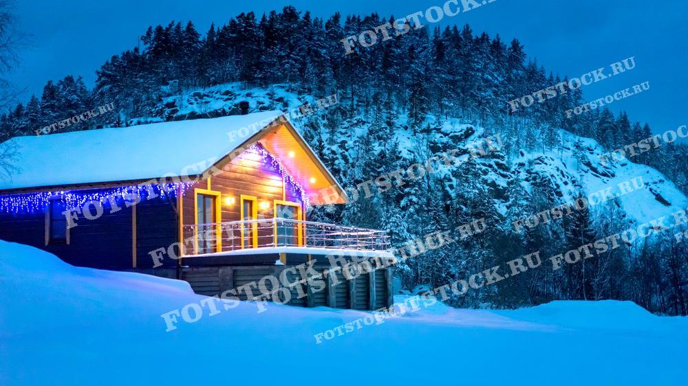 На фотографии изображен загородный дом, украшенный праздничными гирляндами, расположенный на фоне заснеженных гор и леса. Дом светится теплым светом, создавая контраст с холодным зимним пейзажем. Снег покрывает крышу и землю, добавляя ощущение тишины и уединения, что делает это место идеальным для зимнего отдыха.