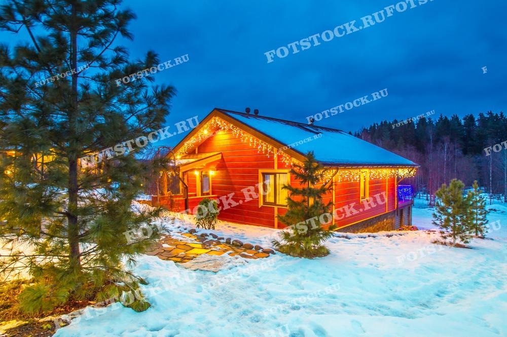 На фотографии изображен загородный дом, украшенный светящимися новогодними гирляндами, который стоит в окружении заснеженного пейзажа. Снег покрывает землю и деревья, создавая атмосферу зимней сказки. Дом светится теплым светом, контрастируя с темным вечерним небом, что усиливает ощущение уюта и праздничного настроения.