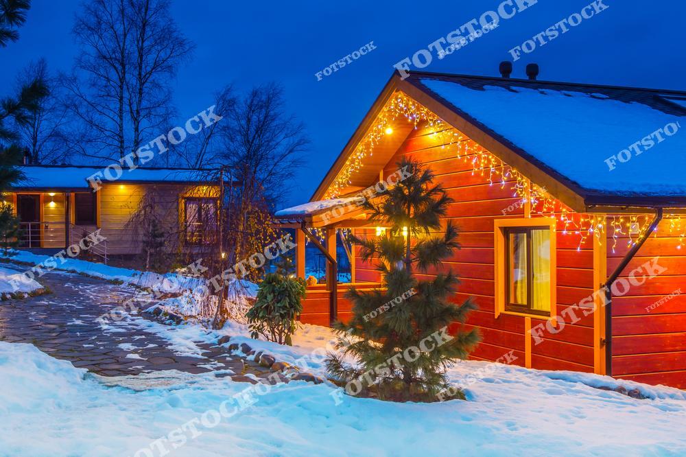 На фотографии изображен загородный дом, украшенный яркими гирляндами, которые придают теплый свет и уютное настроение зимнему вечеру. Снег покрывает землю и крыши, а темное небо создает контраст с праздничной иллюминацией. Этот снимок подчеркивает красоту загородной жизни в зимнее время и уют зимних вечеров.