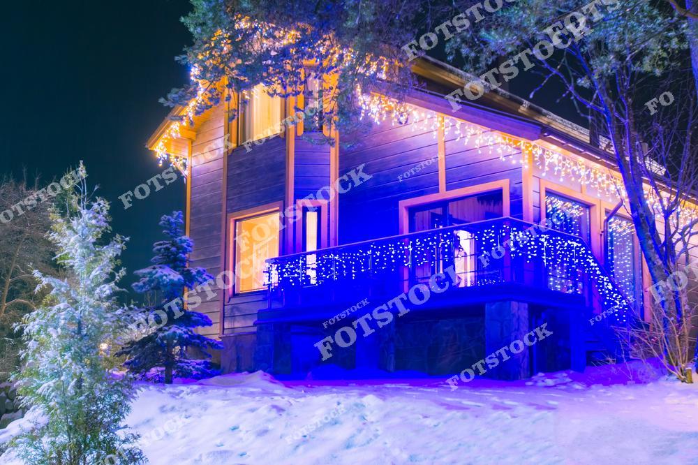 На фотографии изображен загородный дом, украшенный праздничными гирляндами, которые заливают светом фасад и окружающий снежный ландшафт. Яркая подсветка террасы и окон добавляет волшебства зимней ночи. Этот снимок передает атмосферу уюта, праздника и зимнего отдыха.