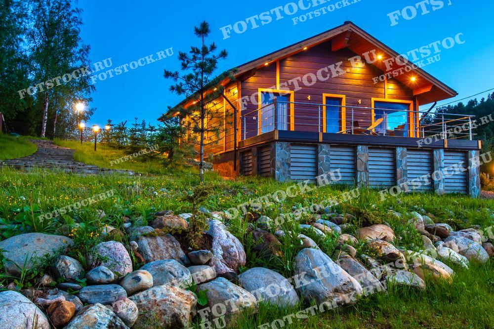 На фотографии изображен загородный дом, расположенный на склоне холма, окруженного зеленью и камнями. Вечернее освещение дома создает теплую и уютную атмосферу. Вокруг дома растут деревья и кустарники, а у подножия холма выложены декоративные камни. Этот дом идеально подходит для уединенного отдыха на природе, вдали от городской суеты.