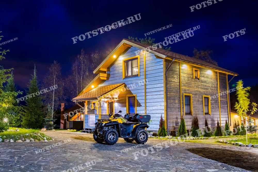 На фотографии изображен загородный дом с вечерней подсветкой, стоящий на ухоженном участке. Перед домом припаркован квадроцикл. Ночная обстановка с подсветкой и окружающей природой подчеркивает уют и комфорт загородной жизни.