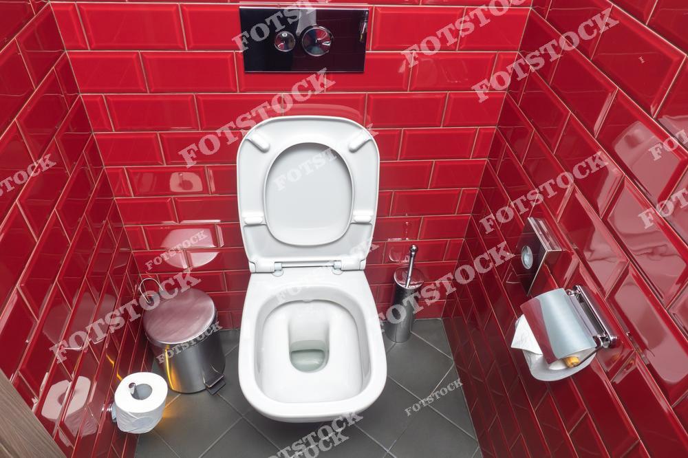 На изображении — современный санузел с белым унитазом, глянцевой красной плиткой на стенах и тёмным кафелем на полу. В интерьере присутствуют элементы гигиены: ершик, мусорное ведро, держатель для туалетной бумаги. Фото отражает яркое дизайнерское решение для небольшой туалетной комнаты.