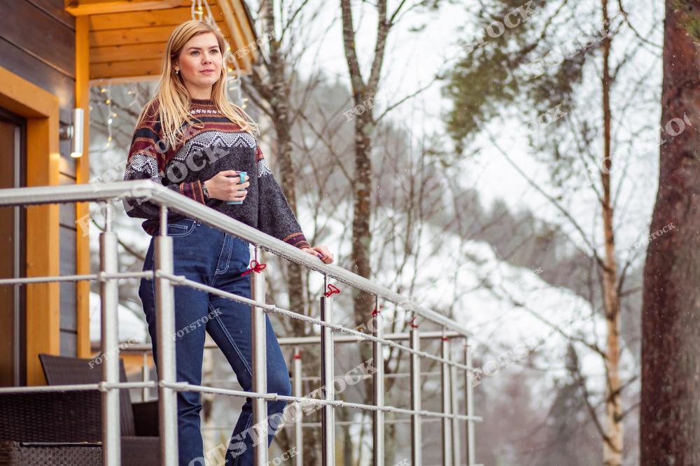 На снимке изображена женщина в тёплом свитере, стоящая на балконе загородного дома с кружкой в руках. На фоне виден зимний пейзаж с деревьями и заснеженными холмами. Её поза и выражение лица передают ощущение уединения и спокойствия.