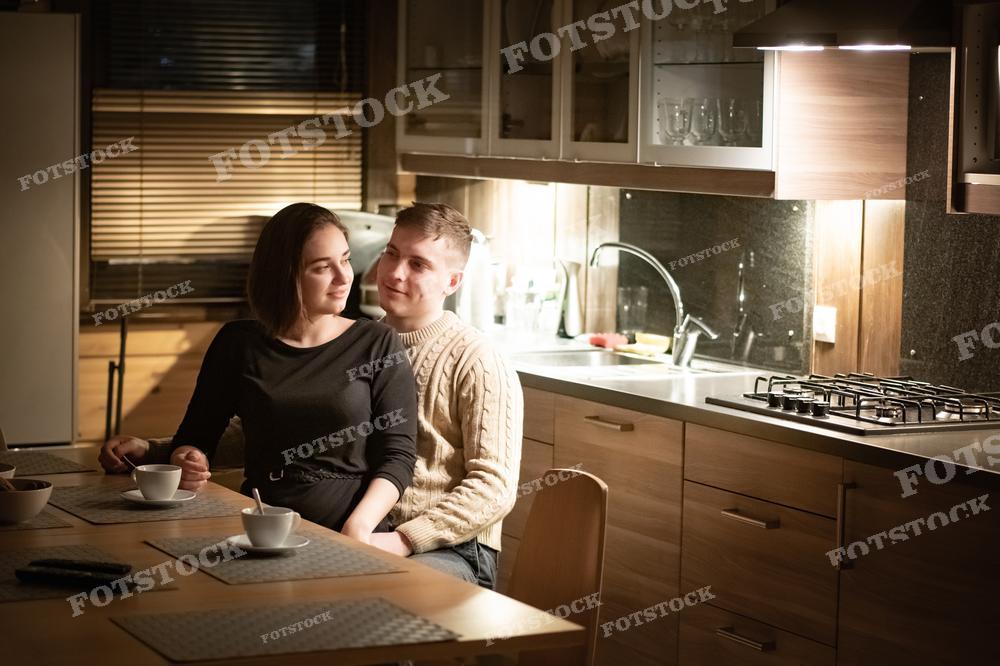 На фотографии изображена молодая пара, уютно расположившаяся на кухне с чашками кофе или чая. Они оба улыбаются и смотрят друг на друга, создавая атмосферу тепла и домашнего уюта. Интерьер кухни выполнен в современном стиле с деревянной мебелью и подсветкой. Фотография может быть использована в статьях о семейных отношениях, дизайне интерьера, в блогах о домашнем уюте, или как рекламный материал для кухонной мебели.