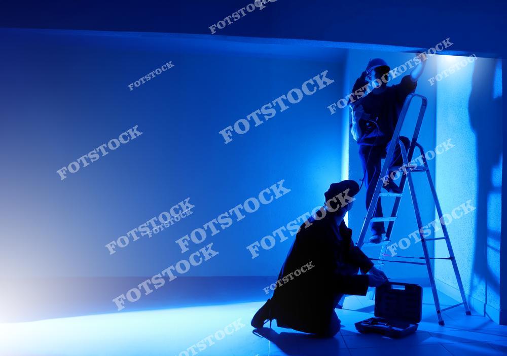 На фотографии изображены два человека в процессе работы над освещением или интерьером в помещении с синим освещением. Один человек стоит на лестнице и что-то регулирует у потолка, второй сидит на полу и работает с инструментами. Оба в рабочей одежде и кепках. Сцена выглядит как подготовка к съемке фильма или настройка осветительного оборудования.