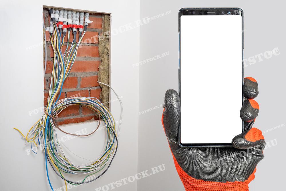 Рука в защитной перчатке держит смартфон на фоне открытой стены с электропроводкой. Этот образ символизирует современный подход к поиску и выбору подрядчика для монтажа электропроводки, включая использование мобильных технологий для оценки, диагностики и мониторинга работ.