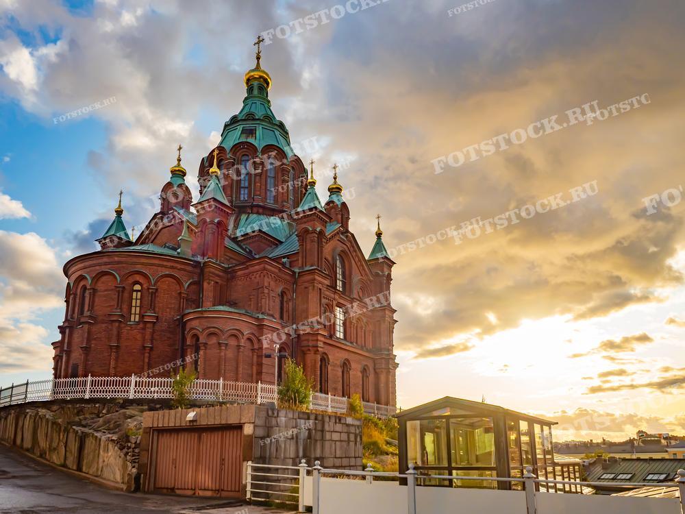 На фотографии изображен Успенский собор в Хельсинки, Финляндия, в летний день. Собор расположен на высоком холме, откуда открывается великолепный вид на город. Это крупнейший православный храм в Западной и Северной Европе, его красные кирпичные стены и золотые купола ярко выделяются на фоне городской застройки и синего летнего неба. Фотография может использоваться для иллюстрации туристических материалов по Хельсинки, промо-материалов туров в Финляндию, а также в образовательных и культурных публикациях, посвященных православной архитектуре.