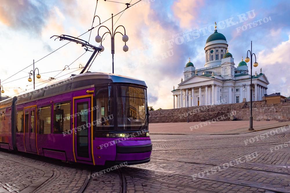 На фотографии представлен красочный вечерний вид на Кафедральный собор Хельсинки, также известный как Собор Святого Николая. На переднем плане изображён фиолетовый трамвай, проезжающий по каменной мостовой, что добавляет сцене динамику и современность.  
Эта фотография отлично подойдёт для публикаций, посвящённых финской архитектуре, городской жизни и транспорту, а также может быть использована в туристических проспектах или как элемент урбанистического дизайна.