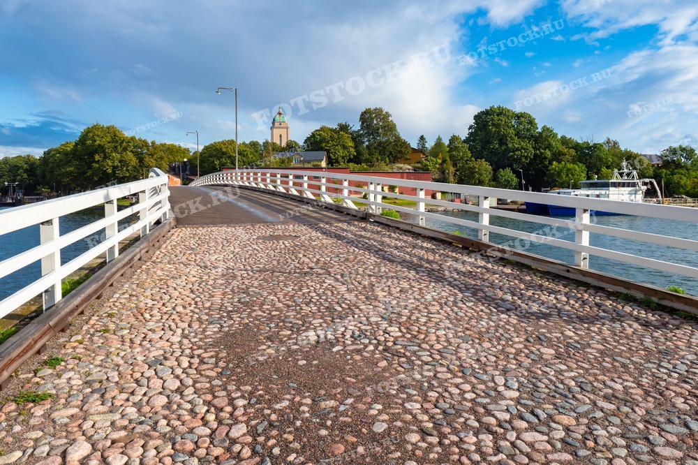 На фотографии — перспективный вид моста, мощёного каменной брусчаткой, который ведёт к зелёному острову с видимым вдалеке колокольным зданием. Небо ясное с лёгкими облаками, что создаёт атмосферу ясного летнего или весеннего дня. Фотография может быть использована в туристических путеводителях, презентациях и публикациях о путешествиях, а также в качестве иллюстрации статей о скандинавской архитектуре и инфраструктуре.