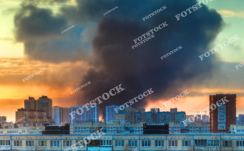 На фотографии изображен городской пейзаж с поднимающимся густым дымом на фоне яркого заката. Черный дым контрастирует с теплым светом, подчеркивая проблемы загрязнения воздуха и экологии.