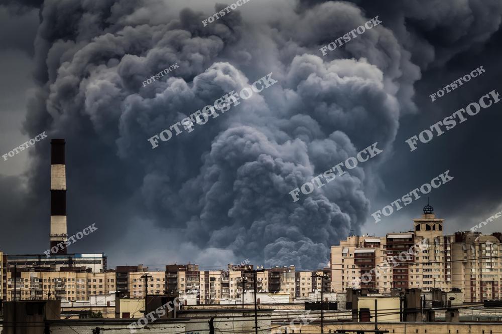 На фотографии изображен городской пейзаж с густыми облаками дыма, поднимающимися над домами. Промышленная труба на фоне усиливает атмосферу экологической проблемы, связанной с загрязнением воздуха и урбанизацией.