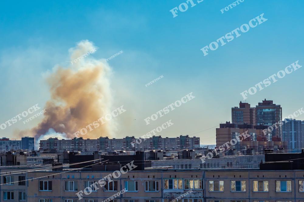 На фотографии изображен столб густого дыма, поднимающегося над жилыми домами в городской черте. Голубое небо контрастирует с желтоватым дымом, подчеркивая серьезность происшествия и его влияние на окружающую среду.