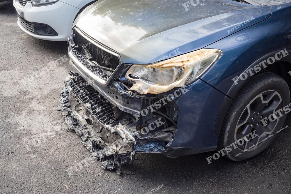 На изображении показан автомобиль с серьёзными повреждениями передней части, вызванными пожаром. Видны обгоревшие и расплавленные элементы бампера и фары. Снимок подчёркивает последствия возгораний в транспортных средствах и важность соблюдения мер предосторожности.