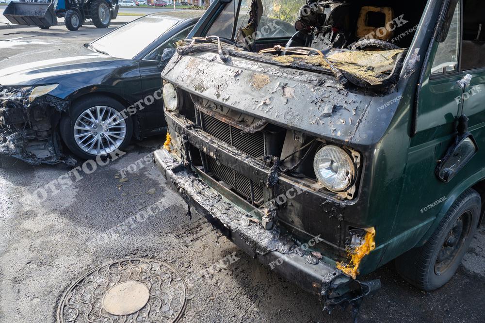На переднем плане изображён микроавтобус с серьёзными повреждениями от огня, включая обугленные и расплавленные элементы. На заднем плане виден легковой автомобиль с аналогичными повреждениями. Снимок подчёркивает серьёзность последствий пожара и актуальность мер безопасности.