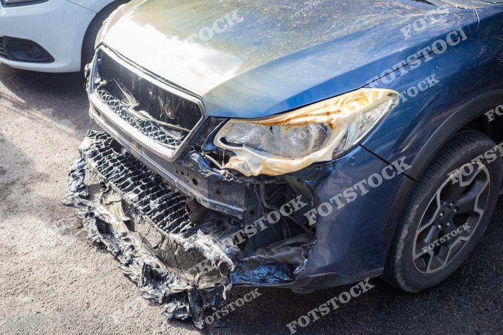 На изображении показан синий легковой автомобиль с серьёзными повреждениями передней части, вызванными пожаром. Расплавленная решётка радиатора и обгоревшая фара подчёркивают разрушительные последствия огня. Фотография акцентирует внимание на необходимости пожарной безопасности для автомобилей.