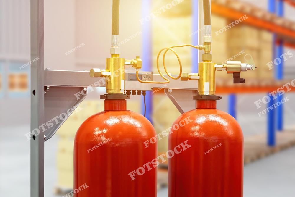 На переднем плане изображены два красных газовых баллона, подключённых к системе управления пожаротушением. На заднем плане видны стеллажи с коробками в складском помещении, подчёркивающие место установки оборудования. Фотография демонстрирует важность современных систем пожаротушения для защиты складских объектов.