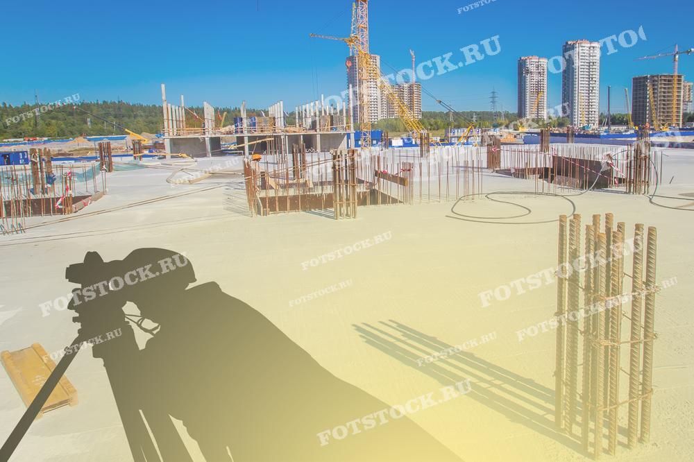 На фотографии изображена тень геодезического нивелира на бетонной поверхности строительной площадки. На заднем плане видны строящиеся здания и башенные краны, что подчеркивает важность геодезических измерений и контроля на различных этапах строительства.