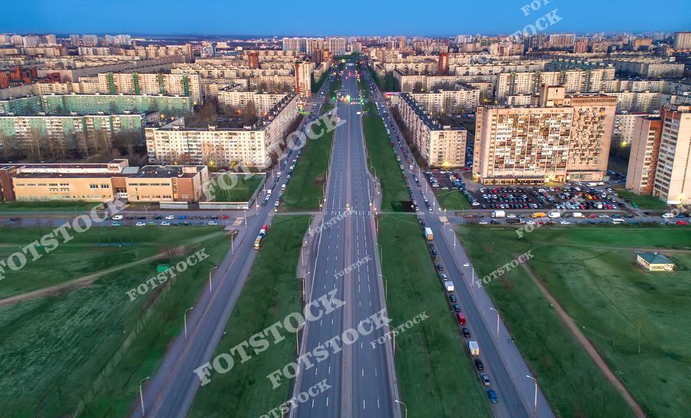 Фотография показывает городской район с многополосной магистралью, окруженной жилыми домами и зелеными участками. Снимок сделан с высоты, подчеркивая масштаб городской инфраструктуры. Вечерний свет добавляет мягкости и уюта к урбанистическому пейзажу.