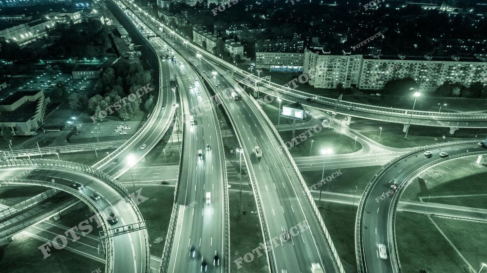На фотографии ночной вид с высоты на разветвленную систему дорожных развязок. Светящиеся огни автомобилей создают динамичные линии на фоне темного неба и окружающей застройки. Освещенные дороги и мосты переплетаются, образуя сложную сеть транспортных артерий.
