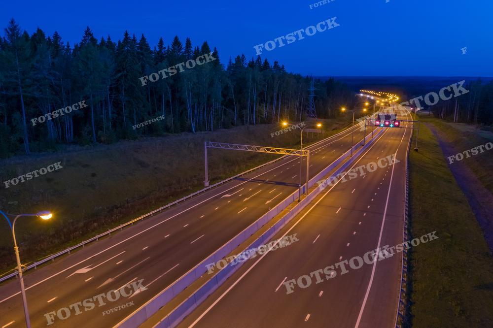 Фотография демонстрирует ночное движение на автостраде, окружённой лесом. Уличные фонари создают ровный ритм света вдоль пути, а дорога тянется вглубь ландшафта, уводя взгляд в тёмное небо. Это изображение может использоваться для представления инфраструктуры и транспорта, в материалах о дорожной безопасности или как фон для различных дизайнерских концепций.