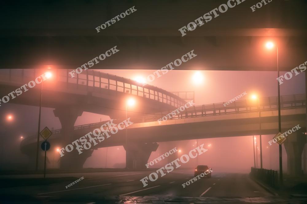 На фотографии изображена мостовая развязка, погруженная в густой туман и освещенная уличными фонарями. Туман и свет создают мистическую и загадочную атмосферу. Снимок подчеркивает сложность и организованность городской транспортной системы.