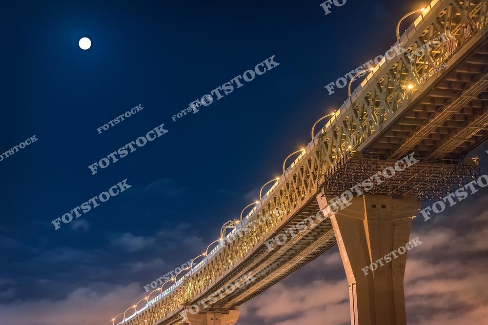 На фотографии изображен фрагмент освещенного моста, снятый ночью на фоне темного неба и полной луны. Конструкция моста подсвечена желтыми огнями, что подчеркивает его детали и современный дизайн. Лунный свет и огни создают атмосферу уюта и величия городской инфраструктуры.