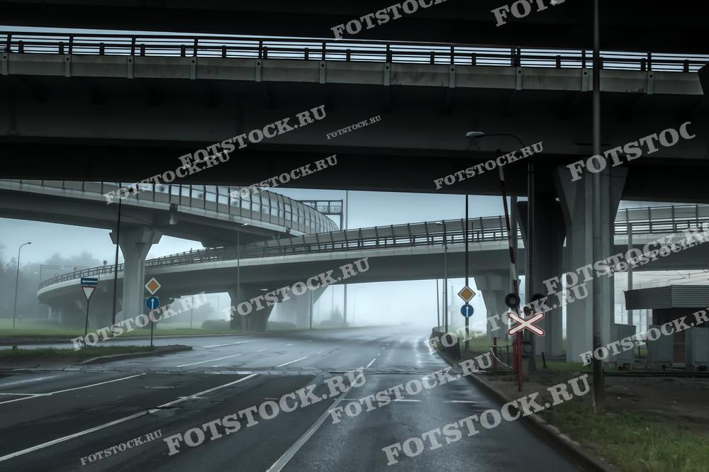 Фотография городской развязки в утренний туман, создающая ощущение пустоты и спокойствия. Снимок подчеркивает особенности городской инфраструктуры в условиях низкой видимости и раннего времени суток.