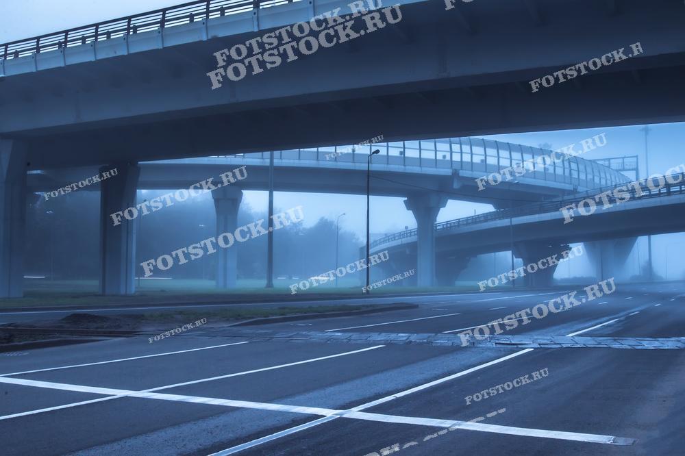 Фотография городской развязки, окутанной густым туманом на рассвете. Снимок передает атмосферу тишины и пустоты, акцентируя внимание на современной городской инфраструктуре и архитектурных линиях.