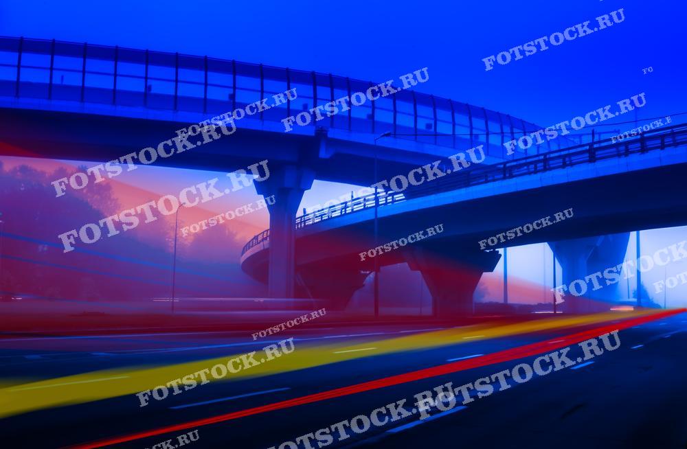 Фотография ночной городской развязки с эффектом размытия световых следов от проезжающих автомобилей. Снимок создает ощущение динамики и движения в атмосфере ночного города, подчеркивая контраст между яркими огнями и темными конструкциями.