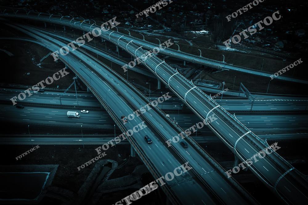 Ночная аэрофотосъемка многополосной автодороги в Петербурге с разделительными полосами и эстакадами. Асфальт отражает мягкий свет фонарей, создавая немного мистическую атмосферу. На дороге видны не многочисленные автомобили, что добавляет ощущение спокойствия и порядка. Цветовая гамма - холодные сине-серые тона.