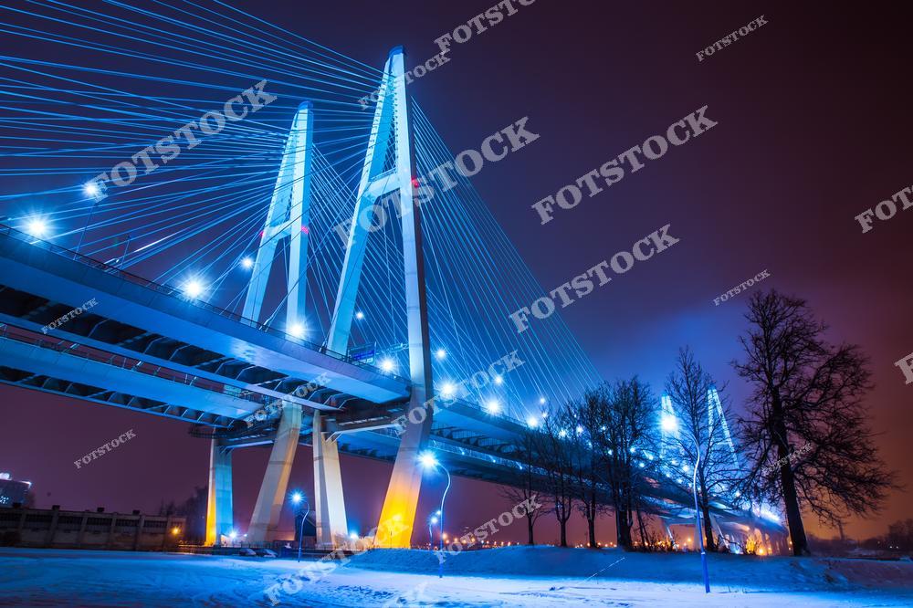 На фотографии изображен ночной канатный мост с яркой подсветкой, пересекающий реку. Световые линии моста создают эффектные визуальные контрасты с темным небом и снежным покровом. Изображение подчеркивает величественную современную архитектуру и атмосферу зимнего города.