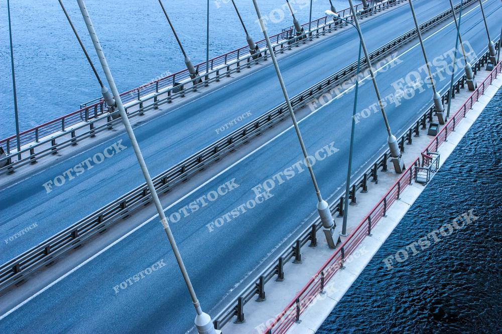 На фотографии изображен подвесной мост, проходящий над голубой водной гладью. Металлические опоры и канаты подчёркивают строгость инженерного дизайна. Вид сверху позволяет оценить масштаб и симметрию конструкции.