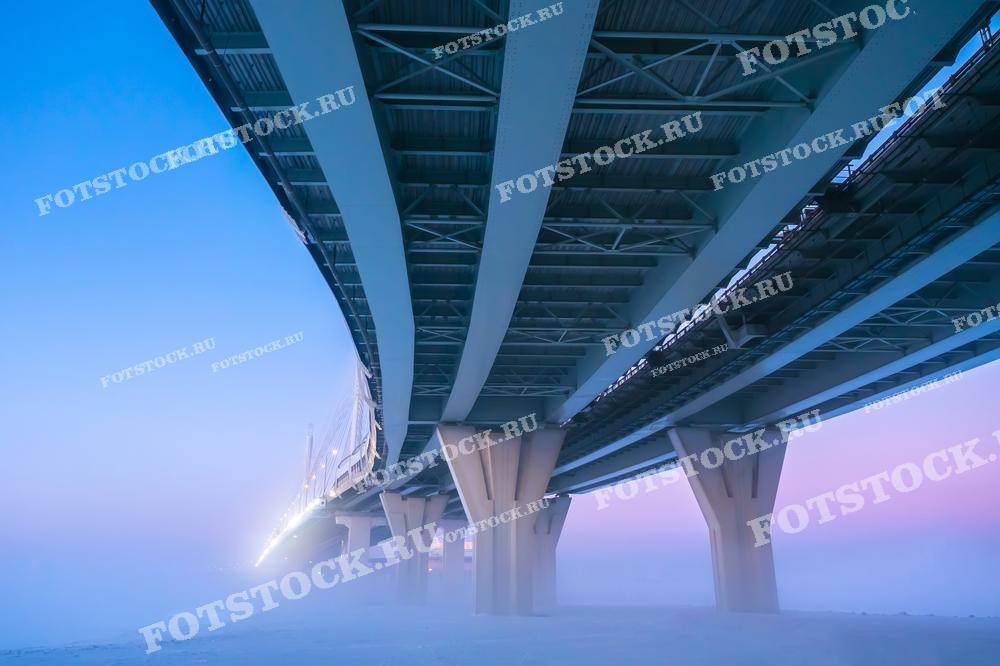 Фотография нижней части моста, простирающегося над туманным ландшафтом, освещенного мягким утренним светом. Снимок подчеркивает масштаб и элегантность архитектурной конструкции в условиях рассветного тумана.