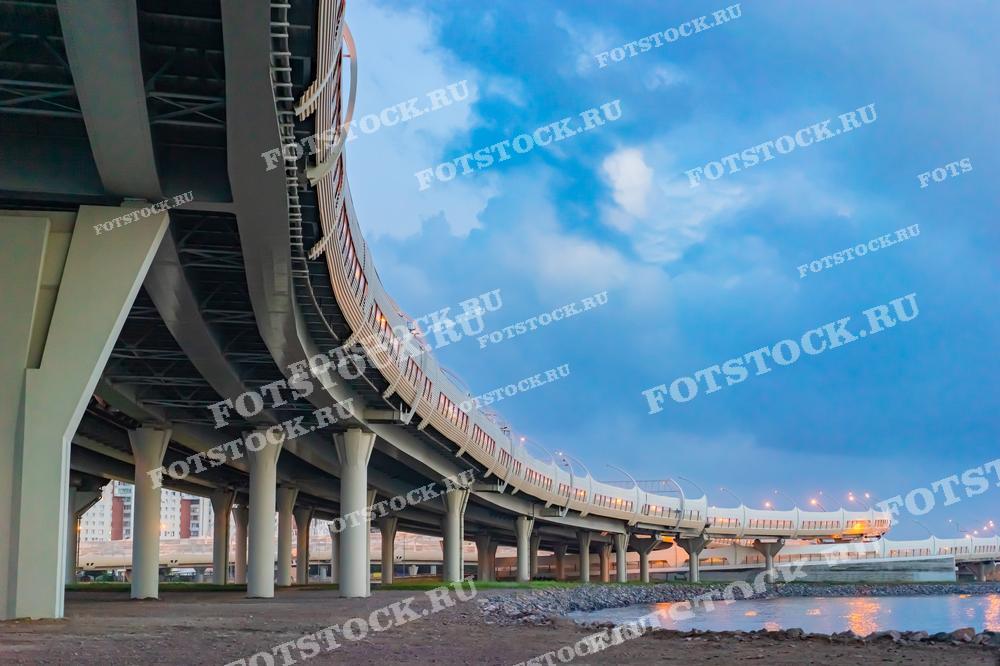 Фотография мостовой развязки, простирающейся над водоемом, на фоне вечернего неба. Снимок подчеркивает современную архитектуру и важность транспортной инфраструктуры в городской среде.