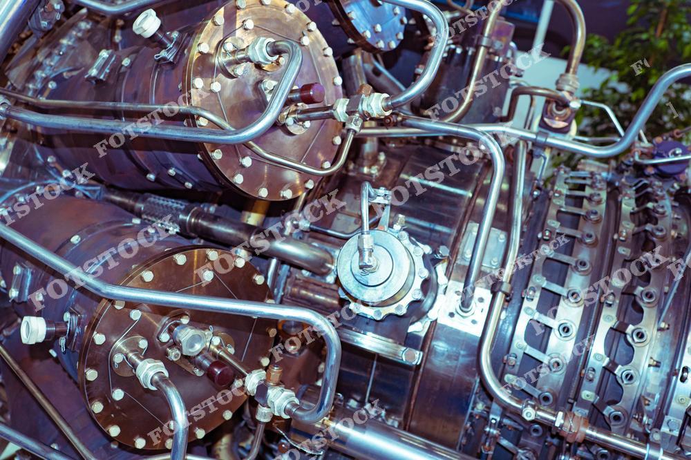 На фотографии показан технически сложный участок газотурбинного или реактивного двигателя. Видны многочисленные металлические трубки, фланцы, болты и системы подачи жидкости или газа. Этот снимок демонстрирует уровень точности и инженерного совершенства в машиностроении, авиастроении или энергетике.