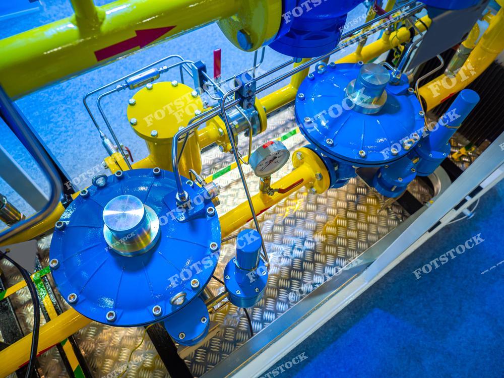 На фотографии изображены газорегуляторные установки с яркими синими и жёлтыми элементами, манометрами и трубопроводами. Снимок символизирует современные технологии контроля и распределения газа в промышленности. Фотография сделана в помещении при ярком освещении на выставочном стенде.