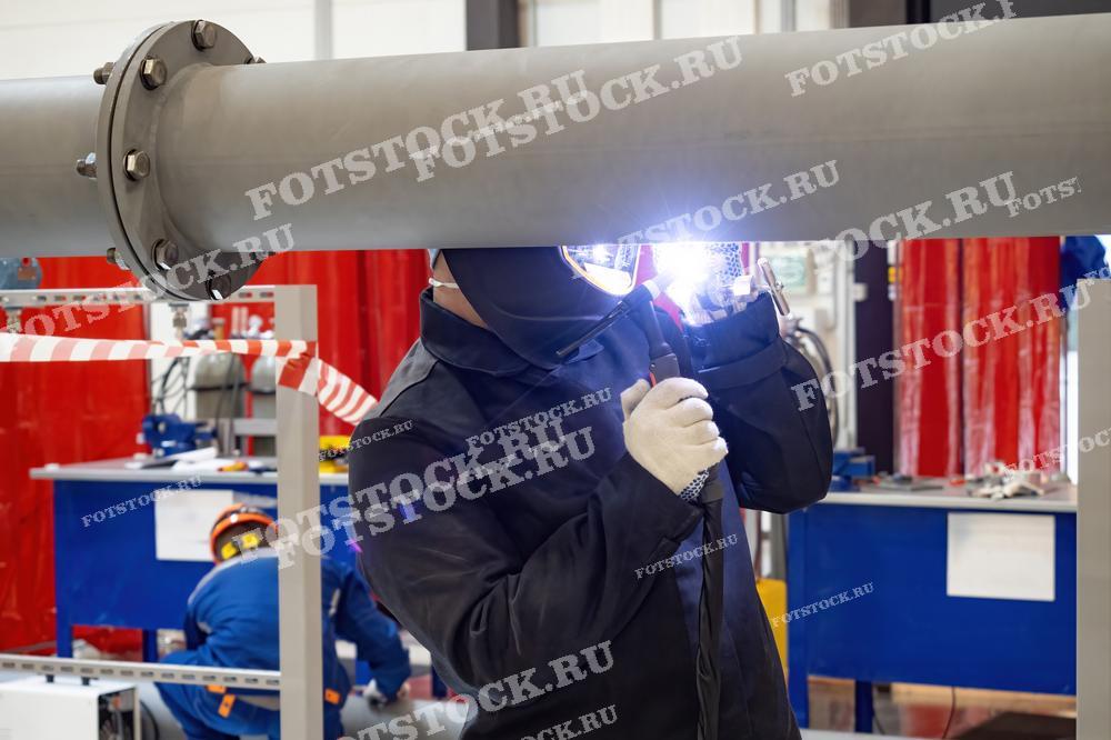 Фотография показывает сварщика в защитной маске и одежде, который выполняет сварочные работы на металлической трубе. На заднем плане видны другие рабочие и оборудование. Это изображение иллюстрирует процесс сварки и монтаж трубопроводов в промышленных условиях.