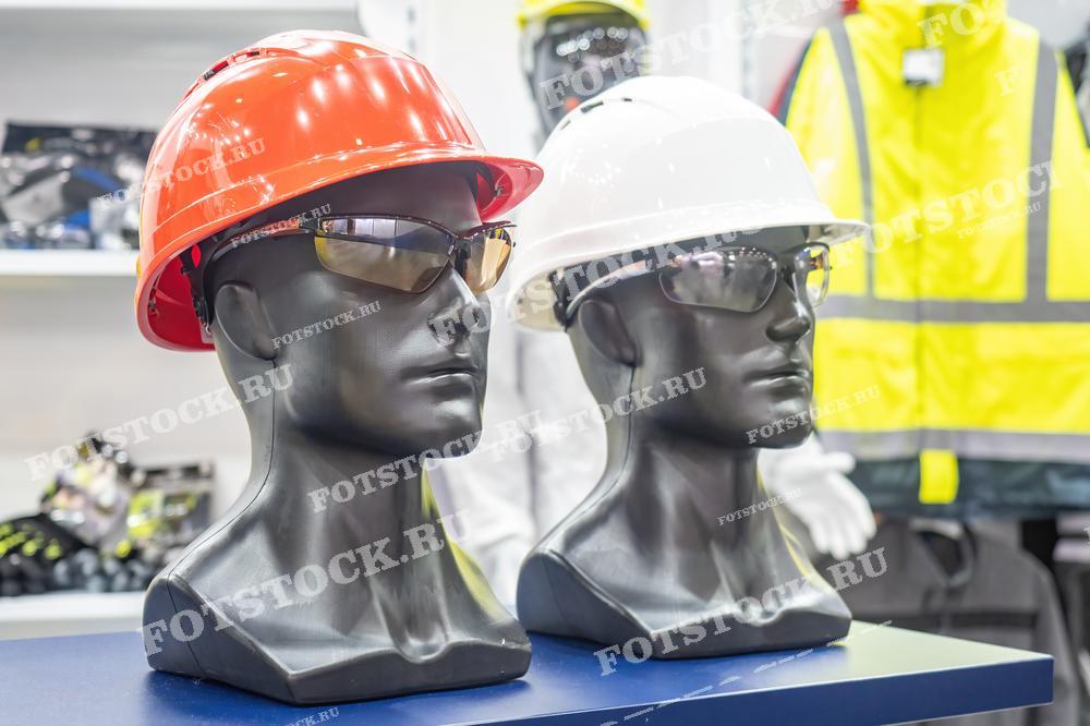 На изображении представлены защитные каски и очки, демонстрируемые на манекенах в выставочном зале. Эти средства индивидуальной защиты предназначены для обеспечения безопасности работников на производстве и строительных объектах.