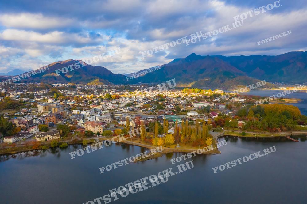 Фотография демонстрирует озеро Кавагутико, расположенное у подножия гор в Японии, с прилегающим к нему городом. Снимок, сделанный с высоты, захватывает осенние краски природы и городской ландшафт, гармонично сочетающиеся с горами на заднем плане.